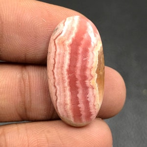 Puede incluir: Una piedra preciosa ovalada en cabujón con rayas rosas y blancas. La piedra tiene una superficie lisa y pulida y un patrón natural y orgánico.
