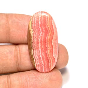 Puede incluir: Una gema ovalada con superficie pulida. La piedra presenta capas de tonos rosa, melocotón y blanco, con una banda marrón en un borde. La gema se sostiene en una mano sobre un fondo blanco.