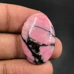 Puede incluir: Una piedra preciosa ovalada en cabujón con un acabado pulido. La piedra es rosa con un patrón de vetas negras en espiral.