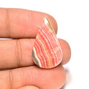 Puede incluir: Una gema en forma de lágrima con un patrón de rayas rosas y blancas, acentuada con tonos beige y grises. La piedra pulida se sostiene en una mano, mostrando su belleza natural y sus variaciones de color únicas. Esta gema probablemente se utiliza para joyería.