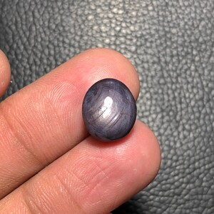 May include: A spherical, polished, dark blue gemstone exhibiting a star effect. The gem has a smooth surface with visible internal striations and a bright, reflective spot. The stone is held between two fingers.