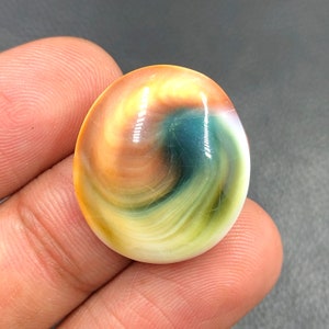 Pode incluir: Uma gema redonda e polida, multicolorida, com padrões de redemoinho de amarelo, laranja, verde e azul. A pedra tem um acabamento liso e brilhante.