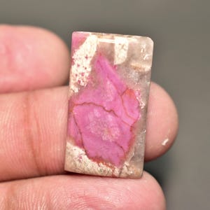 Könnte beinhalten: Ein rechteckiger Edelstein mit einem leuchtend rosa Zentrum und einer durchscheinenden, hellfarbigen Außenschicht. Der rosa Bereich weist ein einzigartiges, strukturiertes Muster mit sichtbaren Rissen auf. Der Stein wird in einer Hand gehalten, was seine polierte Oberfläche zeigt.