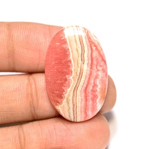Puede incluir: Una gema ovalada con una combinación de colores rosa y crema. La piedra tiene una apariencia en capas con bandas de tonos rosa y crema. La gema se sostiene en una mano.