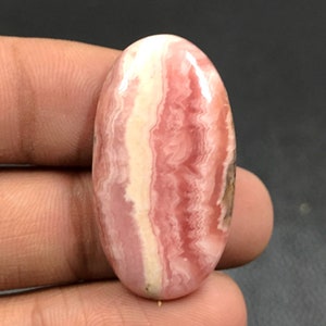 Puede incluir: Una piedra preciosa ovalada en cabujón con un patrón de bandas en tonos rosados y blancos. La piedra está pulida y tiene una superficie lisa.