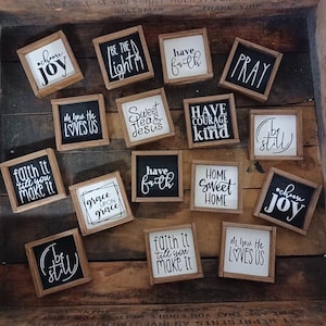 May include: A collection of small, square wooden signs with black and white text. The signs feature inspirational phrases like "Choose Joy," "Be the Light," and "Have Faith." The signs are arranged on a wooden surface.