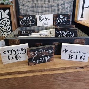 May include: A collection of small wooden signs with inspirational messages, including "Hello beautiful", "Rise above", "Be still Psalm 46:10", "Enjoy life", "Not today Satan", and "Dream big".