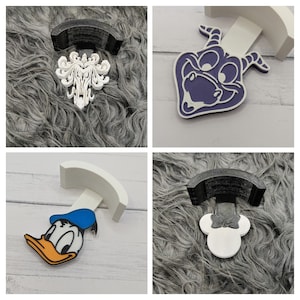 Könnte beinhalten: Vier dekorative Handyhalter mit verschiedenen Designs. Einer hat ein weißes und schwarzes Haunted Mansion-Design, ein anderer eine lila Cartoonfigur, ein dritter Donald Duck und der letzte Minnie Mouse.