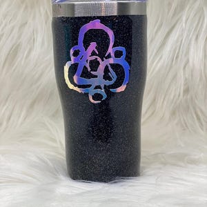 Può includere: Un bicchiere nero con glitter con un logo olografico iridescente di un cerchio con un nodo al suo interno. Il bicchiere ha un coperchio in acciaio inossidabile.