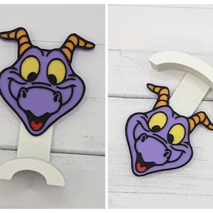 May include: Purple dragon character phone stand with a white base. The dragon has yellow eyes and orange horns.