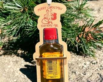 May include: A novelty Christmas ornament featuring a small bottle of Fireball Whisky held in a wooden cutout. The ornament has the text "MERRY CHRISTMAS" and "SHITTER'S FULL". The bottle has a red cap and a yellow label with the text "IGNITE THE NITE".