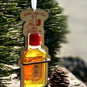May include: A Christmas ornament featuring a small bottle of liquor held within a wooden cutout. The ornament has the text "Merry Christmas" and "Shitter's Full". A pine cone rests nearby, and the ornament hangs from a white ribbon.