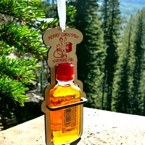 May include: A Christmas ornament featuring a small bottle of liquid held in a wooden frame. The frame has the words "Merry Christmas" and "Shitter's Full" printed on it. The bottle has a red cap and a label that reads "Ignite the Nite".