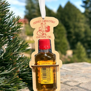 May include: A novelty Christmas ornament featuring a small bottle of Fireball whiskey held within a laser-cut wooden frame. The frame has the words "MERRY CHRISTMAS" and "SHITTER'S FULL" in red. A white ribbon is attached for hanging.
