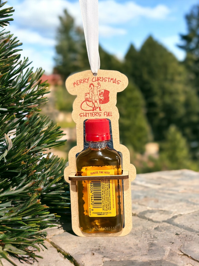 May include: A novelty Christmas ornament featuring a small bottle of Fireball whiskey held within a laser-cut wooden frame. The frame has the words "MERRY CHRISTMAS" and "SHITTER'S FULL" in red. A white ribbon is attached for hanging.