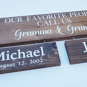 May include: Wooden signs with white lettering. The largest sign reads "OUR FAVORITE PEOPLE CALL US Gramma & Grampa." Two smaller signs below read "Michael August 12, 2002" and "Jessica July 3, 2002."
