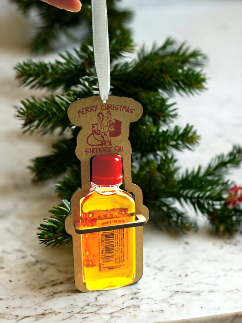 May include: A novelty Christmas ornament featuring a miniature liquor bottle held within a cardboard cutout. The ornament has the text "MERRY CHRISTMAS SHITTER'S FULL" and a red bottle cap. A white ribbon is attached for hanging.
