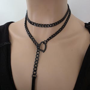 Op de afbeelding: Een zwarte dubbellaagse ketting met een hartvormige sluiting en een verticale rechthoekige staafhanger. De ketting is gemaakt van geschakelde metalen kettingen met een donkere, matte afwerking die op de hals en borst rust.