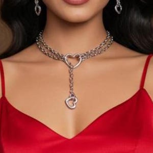May include: A bright red satin dress with thin spaghetti straps and a V-neckline. The outfit is accessorized with a silver-toned chain choker necklace featuring two heart-shaped charms and matching dangling heart-shaped earrings.