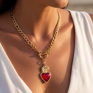 May include: Gold-coloured necklace featuring a red heart pendant. The pendant has a heart-shaped red stone with a gold flame design. The necklace has a thick chain and a toggle clasp. The person is wearing a white dress.