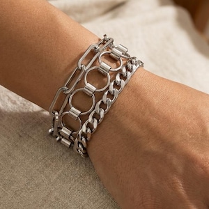May include: A multi-strand silver-toned bracelet set featuring three distinct chain styles. The design includes a rectangular link chain, a central row of interconnected circular rings, and a classic curb chain, all crafted with a polished metallic finish.