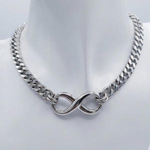 May include: Silver-toned necklace with a curb chain design. The necklace features an infinity symbol as a central pendant. The jewelry is displayed on a white mannequin bust, showcasing the necklace's design and style.