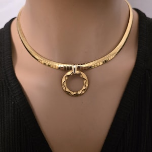 Puede incluir: Un collar dorado con un colgante circular texturizado. El collar tiene un diseño acanalado y un pequeño elemento decorativo en la parte superior del colgante. El colgante tiene un borde ondulado.