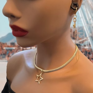 Könnte beinhalten: Eine goldfarbene Halskette mit Sternanhänger und passenden Ohrringen. Die Ohrringe haben ein Stern- und Ketten-Design. Der Schmuck wird auf einer Schaufensterpuppe mit rotem Lippenstift und einem verschwommenen Hintergrund getragen.