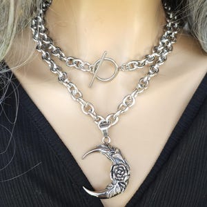 May include: A silver chain necklace with a large, ornate crescent moon pendant featuring a rose design. The necklace is layered with two chains, one with a toggle clasp.