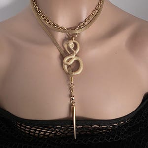 May include: A gold-toned multi-layered necklace featuring a thick chain and a flat mesh band. A textured snake pendant is coiled in the centre, connected to a long, slender, pointed spike charm that hangs vertically from the snake's base.