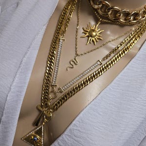 Peut inclure: Plusieurs colliers dorés superposés, comprenant un pendentif soleil, un serpent et une croix avec un symbole d'œil. Les chaînes sont de différents styles et épaisseurs, créant un look audacieux et tendance.
