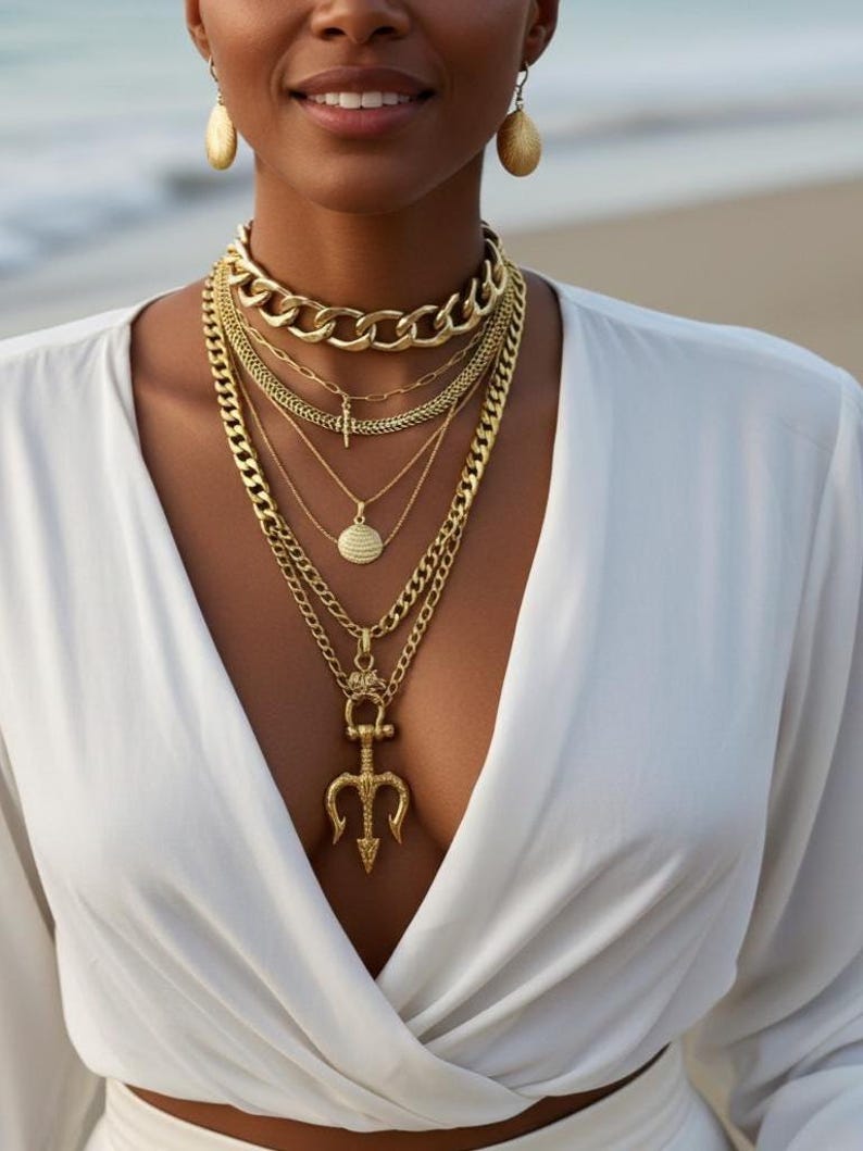 May include: A collection of gold necklaces and earrings. The necklaces include a chunky chain choker, delicate chains, and a pendant with a trident design. The earrings are teardrop-shaped. The jewelry is worn with a white top.