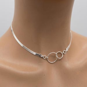 May include: A silver herringbone chain necklace featuring two interlocking circular rings as a central focal point. The necklace is crafted from polished sterling silver, with a small 925 hallmark visible on the connector link near the rings.
