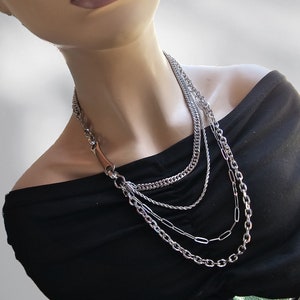 May include: A layered silver chain necklace with a unique, slightly curved, silver pendant. The necklace is worn on a mannequin with a black off-the-shoulder top.