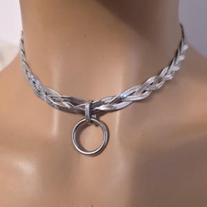 Op de afbeelding: Een zilverkleurige choker-halsketting met een gevlochten visgraatkettingontwerp. De ketting is voorzien van een centrale ronde metalen ringhanger die aan de gevlochten strengen is bevestigd, wat zorgt voor een samenhangende metalen uitstraling.