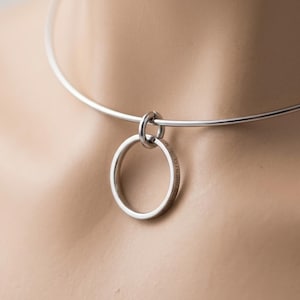 May include: A silver-toned metal choker necklace featuring a thin, circular band and a single, larger circular pendant ring attached by a small jump ring. The pendant ring is engraved with the text, "EVEN WHEN I CLOSE MY EYES."