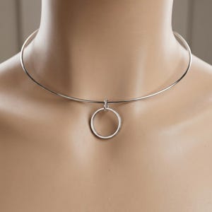 May include: A silver-coloured metal choker necklace featuring a thin, circular wire band that sits close to the neck. A single, smooth, circular ring pendant is attached to the centre of the wire band by a small, integrated jump ring connector.
