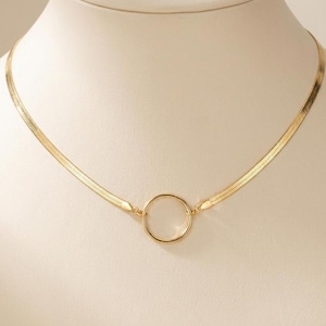 Op de afbeelding: Een goudkleurige ketting met een cirkelvormige hanger. De ketting heeft een plat visgraatpatroon. De hanger is een eenvoudige, open cirkel. De ketting wordt gepresenteerd op een witte mannequin.