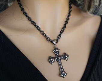 Könnte beinhalten: Eine schwarze Kettenkette mit einem silbernen Kreuzanhänger. Das Kreuz hat ein detailliertes Design mit kleinen silbernen Nieten und einer schwarzen Oberfläche.