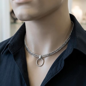 May include: A silver-colored chain necklace with a large, round pendant. The necklace is made of thick, interlocking links and is displayed on a mannequin wearing a black collared shirt. The pendant hangs from a small loop attached to the chain.