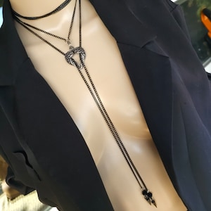 May include: A layered black chain necklace with a silver pendant shaped like a crescent moon. The pendant has a textured design and the necklace has a long, dangling chain with two small black beads at the end.