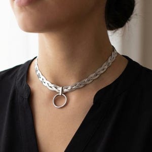 May include: A silver necklace featuring a braided design and a circular pendant. The choker-style necklace is worn against a black top. The necklace is a stylish piece of jewellery.