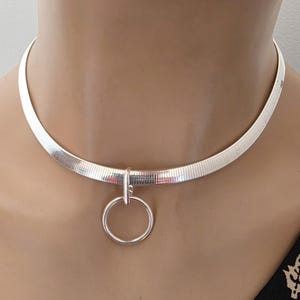 May include: A silver-toned herringbone chain choker necklace featuring a smooth, polished circular ring pendant attached to the center. The necklace has a flat, flexible link design that sits closely around the neck, creating a sleek, metallic appearance.