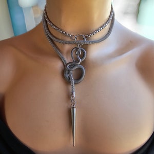May include: A silver-toned necklace with a layered design. The top layer is a chain, the middle layer has a circular design, and the bottom layer features a snake-like design with a long, pointed pendant. The necklace is displayed on a mannequin.