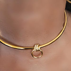 May include: A gold-toned metal choker necklace featuring a smooth, rigid circular band. The center of the necklace is adorned with a decorative knot element that holds a small, circular ring pendant, creating a polished and reflective metallic finish.