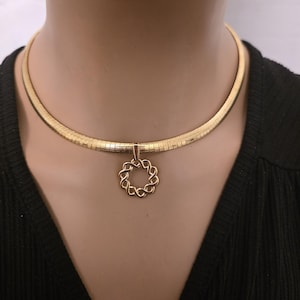 May include: A gold-colored necklace featuring a thick, ribbed choker-style chain. A delicate, openwork pendant in a circular design hangs from the center. The necklace is displayed against a black top.
