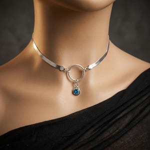 May include: A silver-toned herringbone chain choker necklace featuring a central circular ring connector. A small infinity-shaped charm hangs from the ring, set with a single round, faceted blue gemstone at its base, resting against the neck.