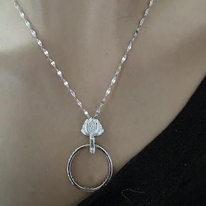 May include: A silver-toned necklace featuring a delicate chain with reflective links and a pendant consisting of a small, textured floral-shaped bail attached to a smooth, circular ring. The metal has a bright, polished finish throughout the piece.