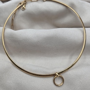 May include: A gold-toned metal choker necklace featuring a smooth, rigid circular band. A small, matching gold-toned ring charm is suspended from the bottom center of the band. The necklace includes a thin chain extender with a lobster clasp.