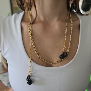 May include: Gold-tone chain necklace with black accents. The necklace features two different chain styles and several black, circular pendants. The necklace is worn over a white top.
